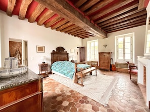 A bedroom with a bed, wooden furniture, a stone floor, and a red-beamed ceiling.