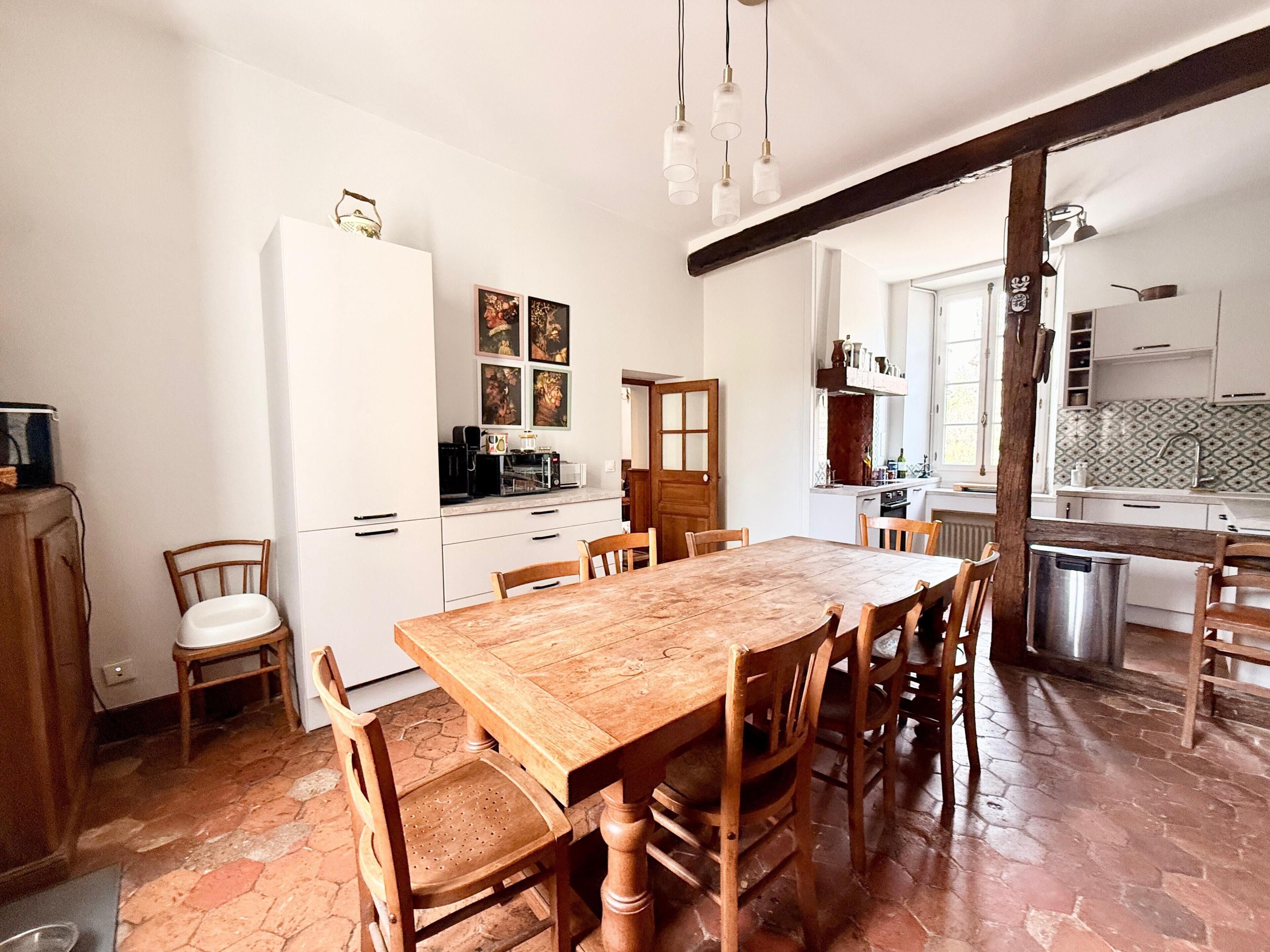 A dining area with a wooden table and chairs, a white refrigerator, and a kitchen in the background.