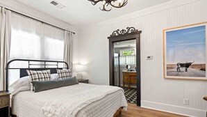 Une chambre à coucher comprenant un lit, une table de chevet, une commode, un miroir, une porte donnant sur une salle de bain et un tableau encadré accroché au mur.