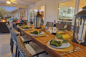 An outdoor dining area with wicker furniture, a long wooden table set for a meal, and a view of the kitchen through large windows. - Enchanted Vine - Pool, Spa, Sauna & Speakeasy (Fredericksburg)