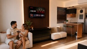 En moderne stue med sofa, et fjernsyn der viser et program, et køkkenområde og et bakke med mad og drikke.