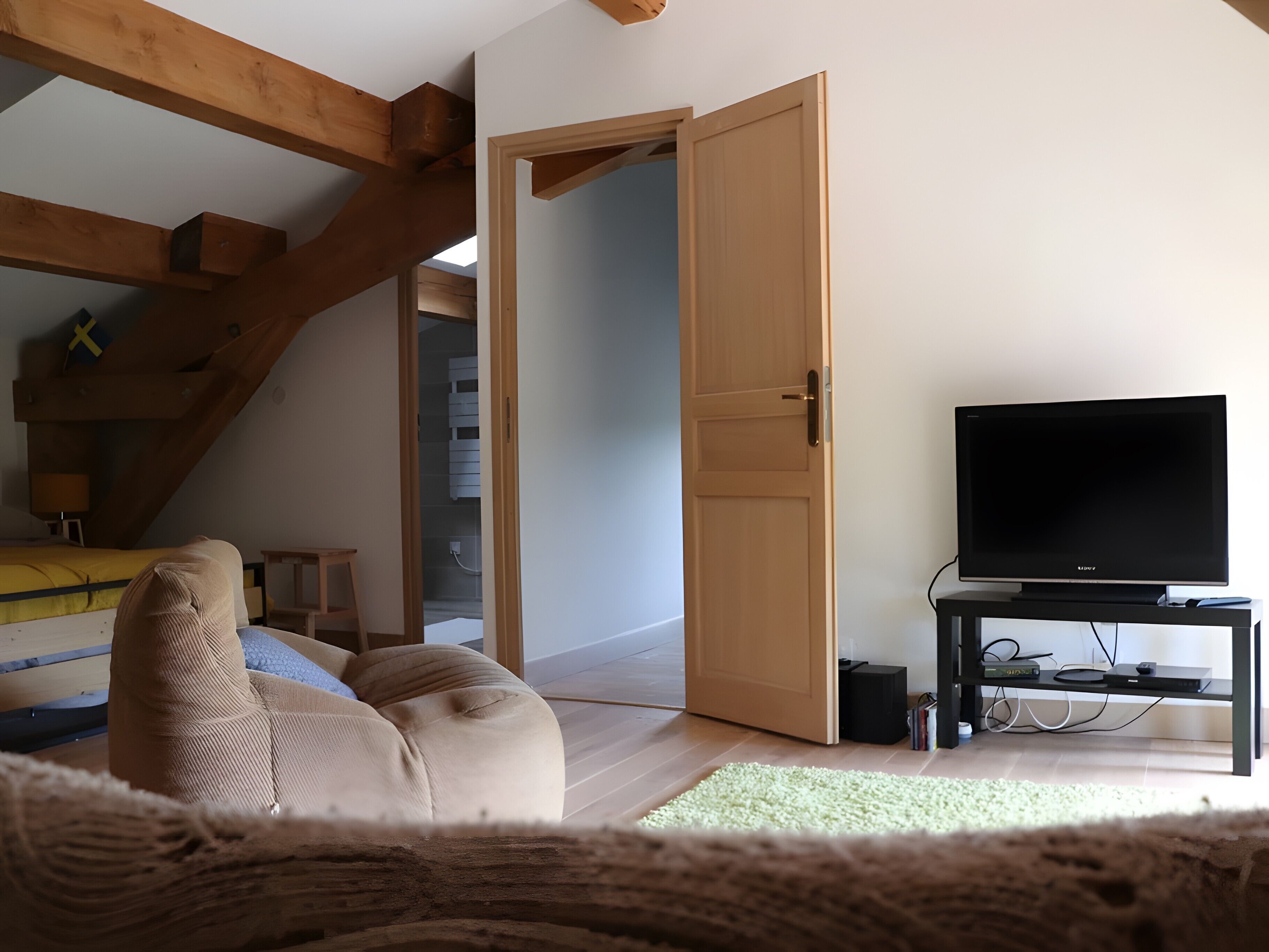 Una habitación con una cama, un televisor sobre un mueble bajo, techo de madera y una puerta que da a otra habitación.
