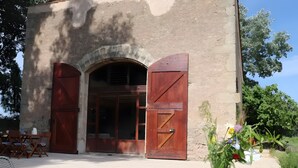 Un bâtiment rustique doté d’une grande porte en arc de cercle, de volets en bois et d’une table sur laquelle se trouvent un pichet jaune, une tasse blanche et un vase rempli de fleurs.