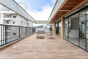 Un balcón amplio con techo de madera, una banca y una silla, con vistas a un edificio de varios pisos y una montaña al fondo.