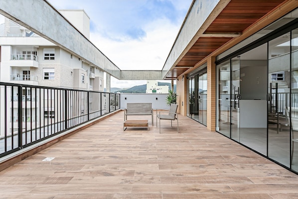 Ein geräumiger Balkon mit Holzboden, einer Bank und einem Stuhl, mit Blick auf ein mehrgeschossiges Gebäude und einen Berg im Hintergrund.