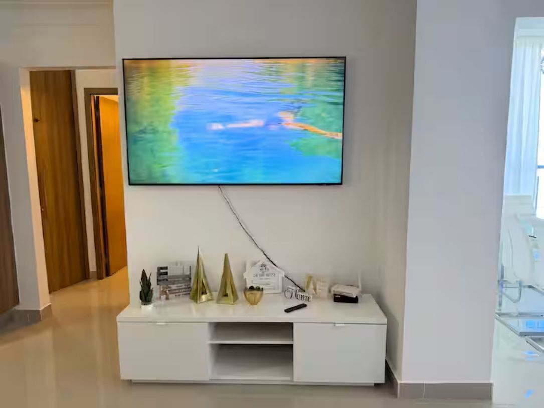 Una sala de estar moderna con un televisor de pantalla plana montado en la pared, un mueble blanco de entretenimiento debajo y una puerta de madera al fondo.