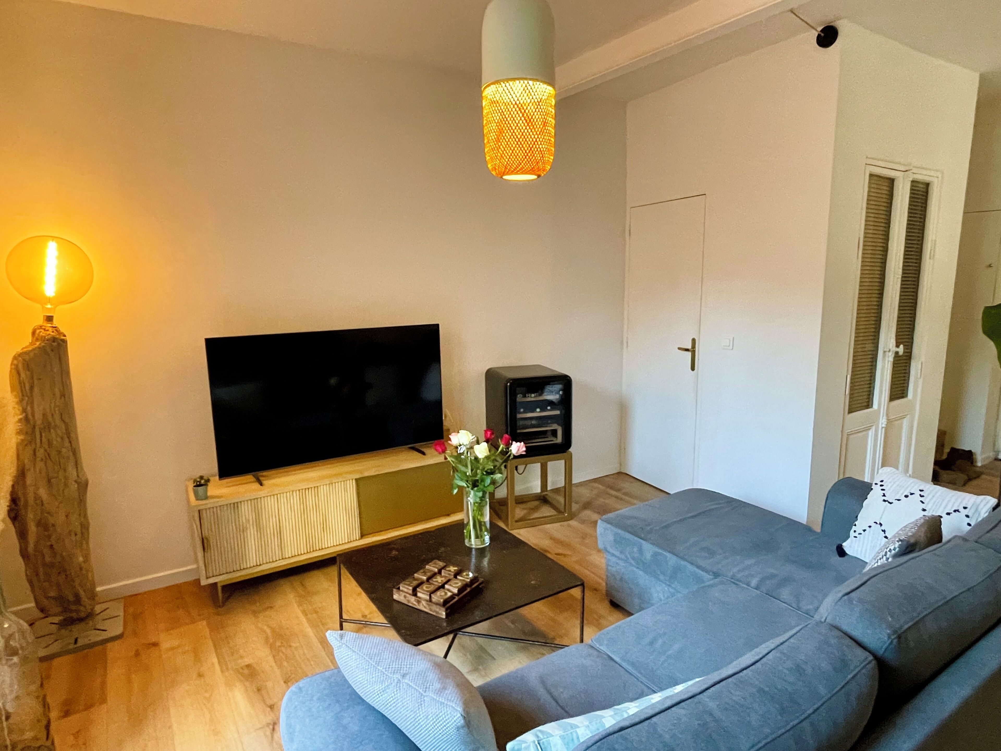 A living room with a flat-screen TV, a coffee table with a vase of flowers, a grey sofa, and a hanging light fixture.