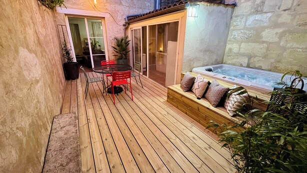 Une terrasse en bois avec un jacuzzi entouré de parois en verre, des chaises rouges et une table.