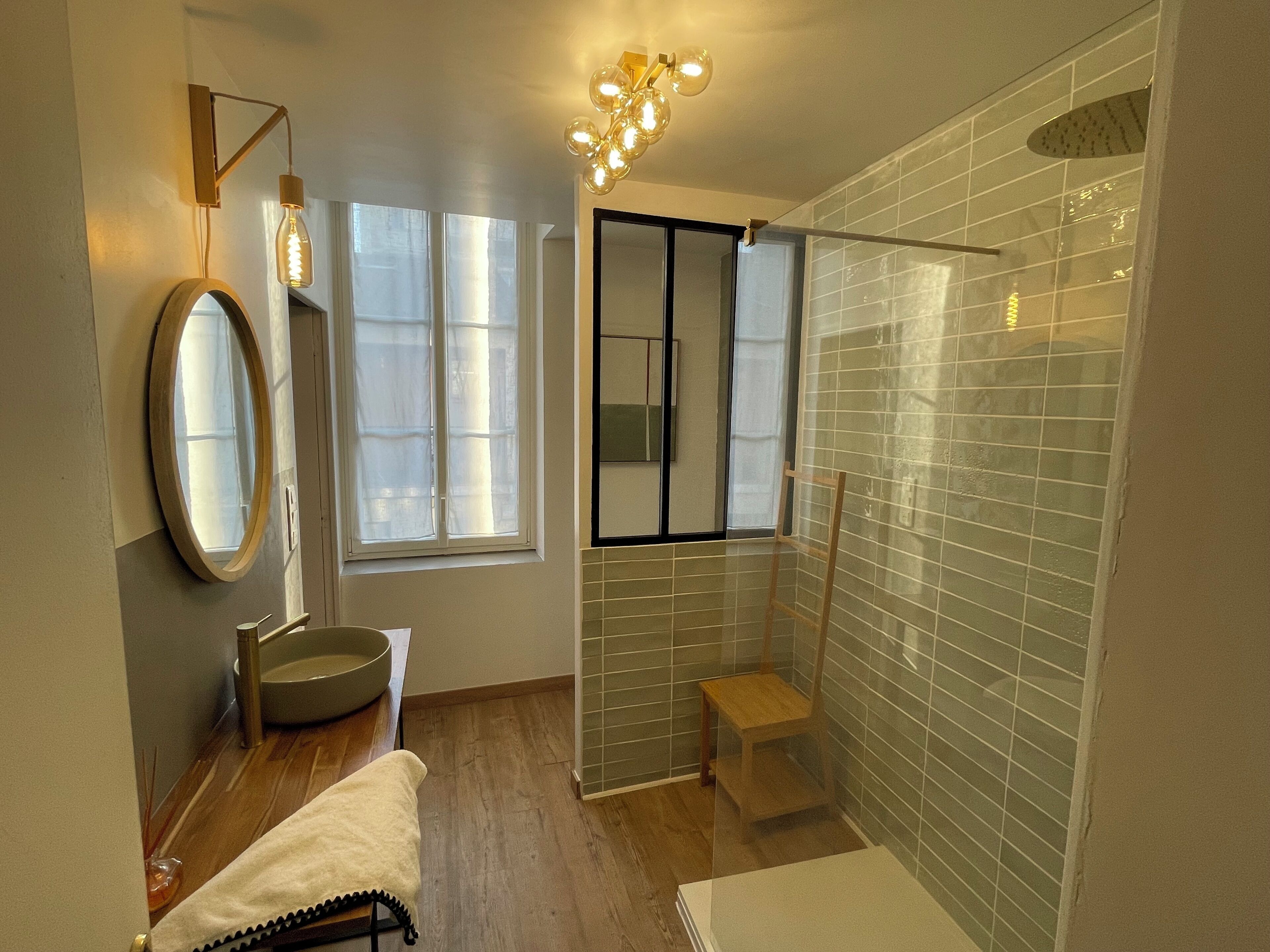 A modern bathroom with a large mirror, a round sink, a shower with a glass door, and a wooden chair.