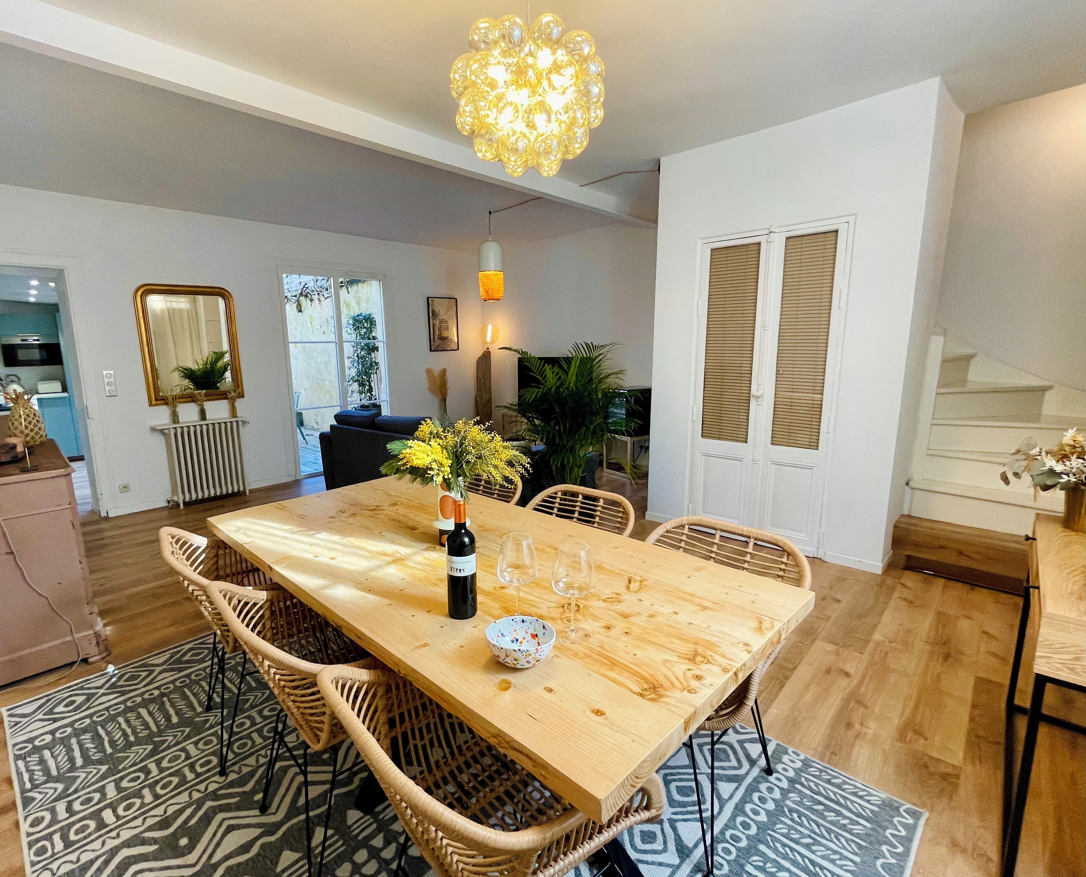 A dining area with a wooden table, wicker chairs, a vase with flowers, and a hanging light fixture.