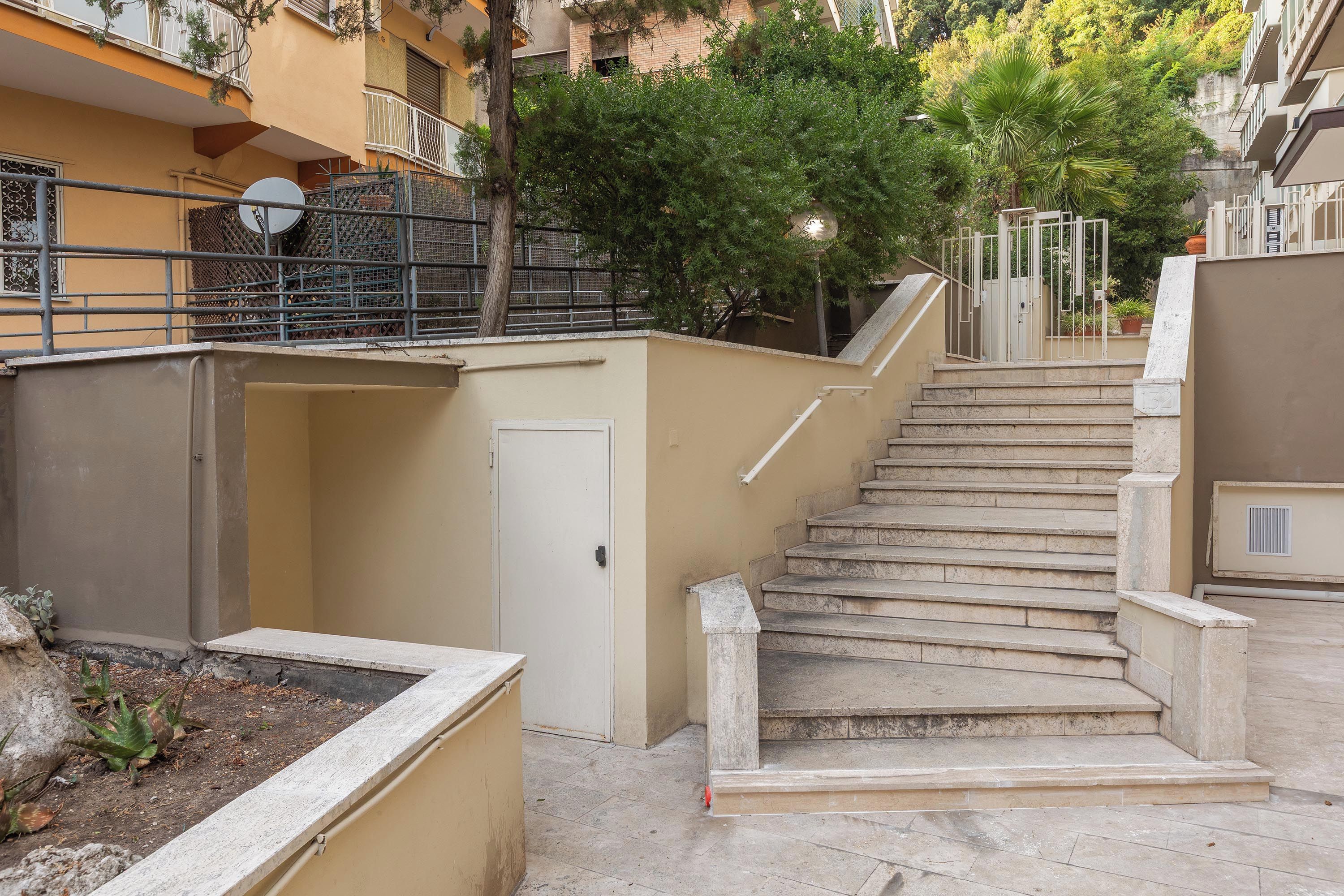 Unos escalones de piedra conducen a la entrada de un edificio, con una puerta y una pequeña zona ajardinada a la izquierda.