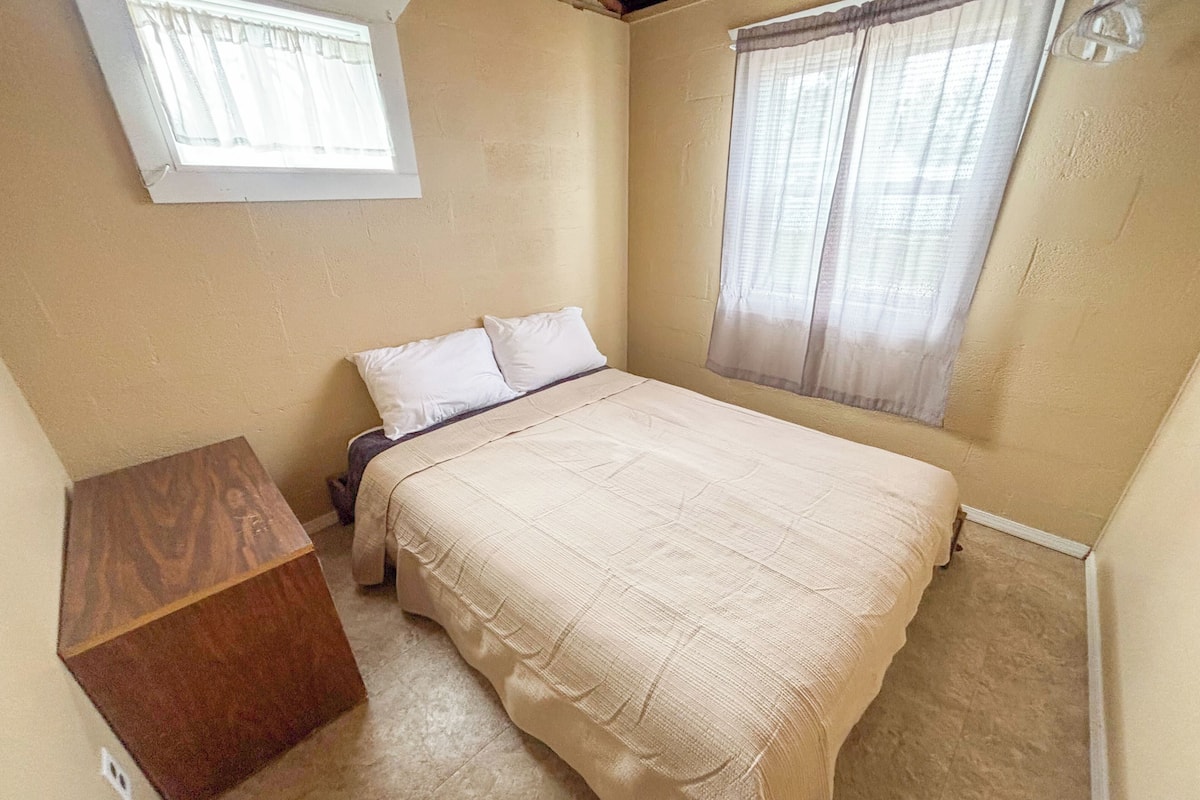 A bedroom with a bed, a nightstand, a wooden cabinet, and two windows with curtains.