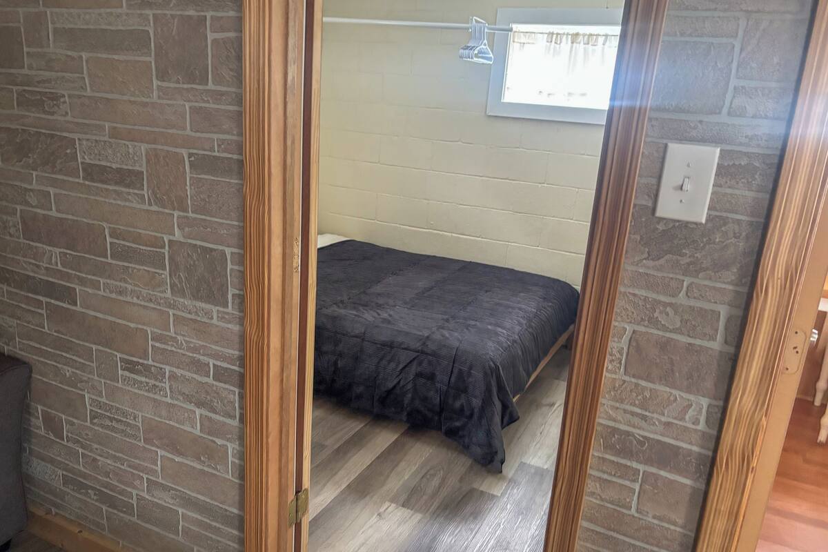A room with a brick wall, a bed with a dark bedspread, a window with curtains, and a light switch on the wall.