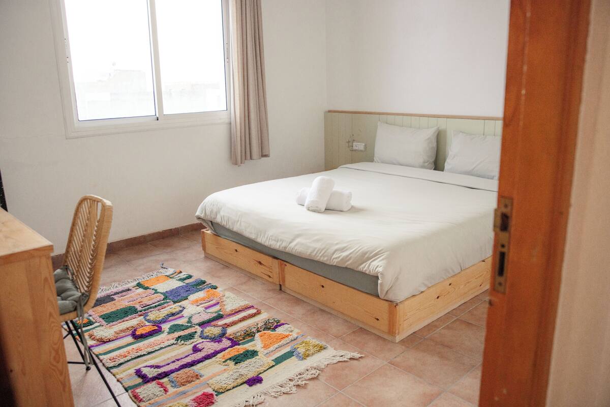 Ein Schlafzimmer mit einem Bett, einem Sessel, einem farbenfrohen Teppich und einem Fenster mit Vorhängen.