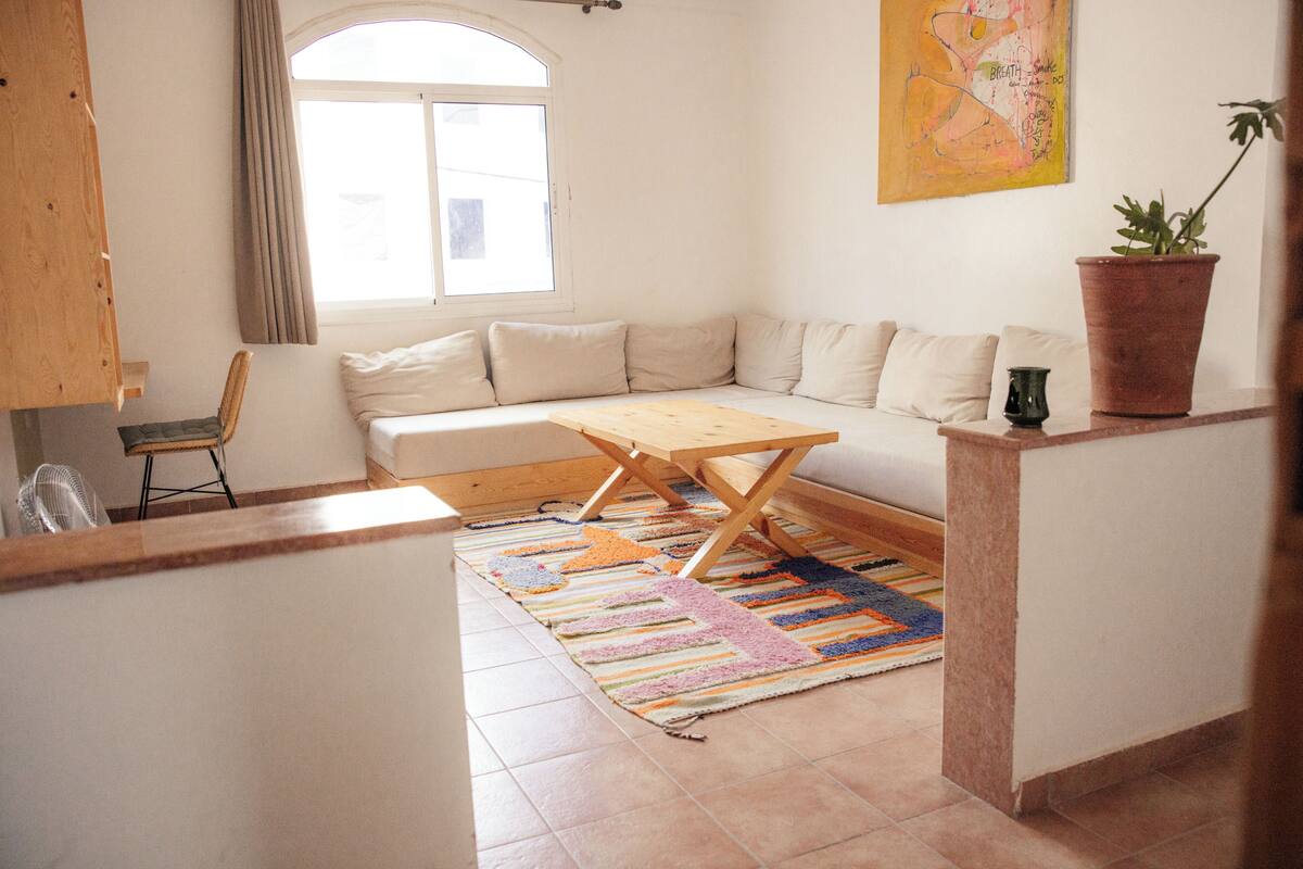 Ein Zimmer mit einer Couch, einem Holztisch, einem Ventilator, einem Fenster mit Vorhängen, einem bunten Teppich und einer Topfpflanze.