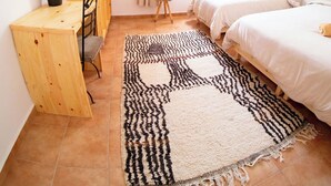 Un dormitorio con dos camas, un escritorio de madera, una silla, un taburete pequeño, una planta en maceta, una alfombra estampada y una ventana con cortinas.