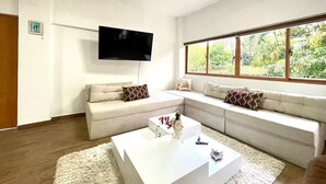 Ein modernes Wohnzimmer mit einem Ecksofa, einem Flachbildfernseher und einem großen Fenster mit Blick auf Grünflächen.
