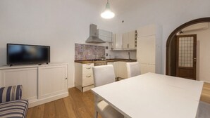 Cocina compacta con gabinetes blancos, protector contra salpicaduras de azulejos, una mesa de comedor y un televisor.
