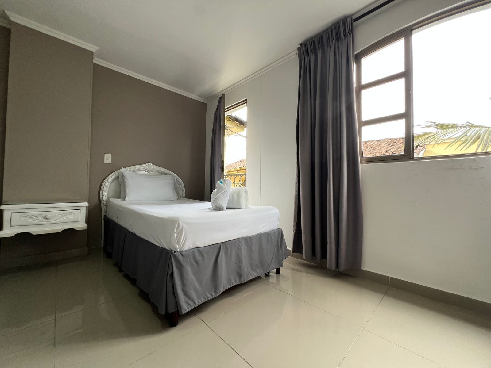 A hotel room with a bed, a bedside table, a window with curtains, and a view of palm trees.