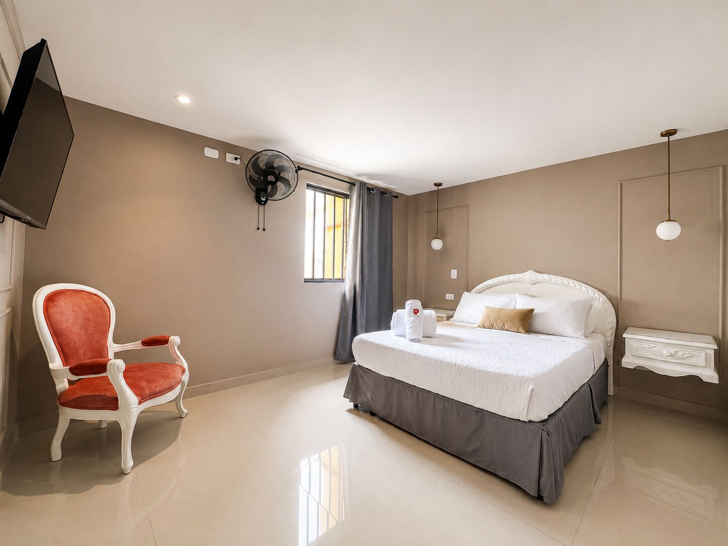 A hotel room with a bed, a chair, a TV, a fan, and a window.