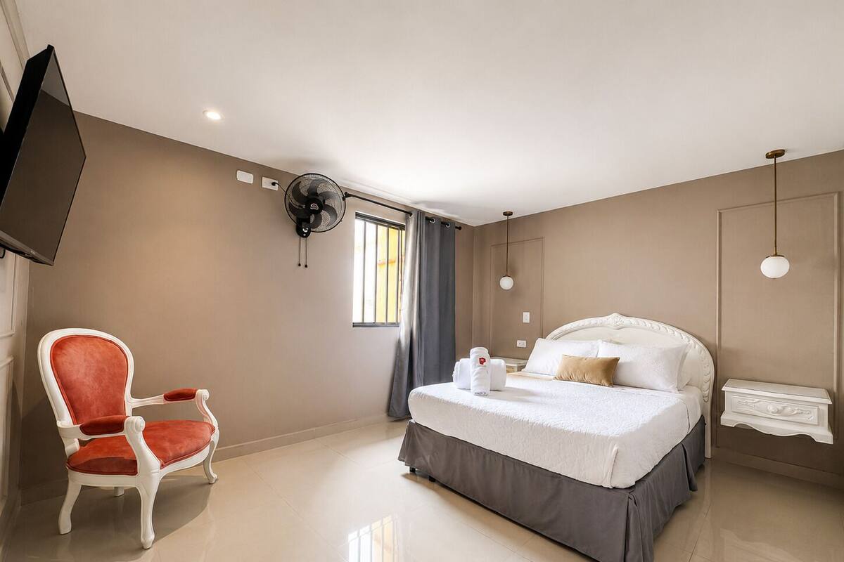 Una habitación de hotel con una cama, una silla, un televisor, un ventilador y una ventana.