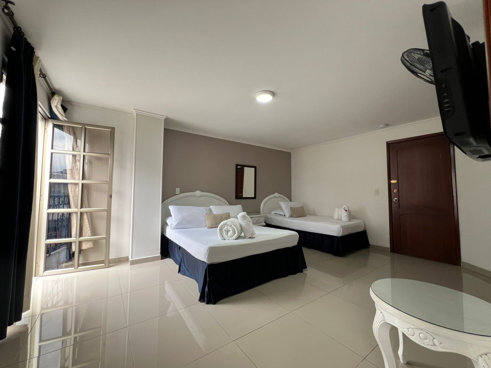 A hotel room with two beds, a television, a glass table, and a door.