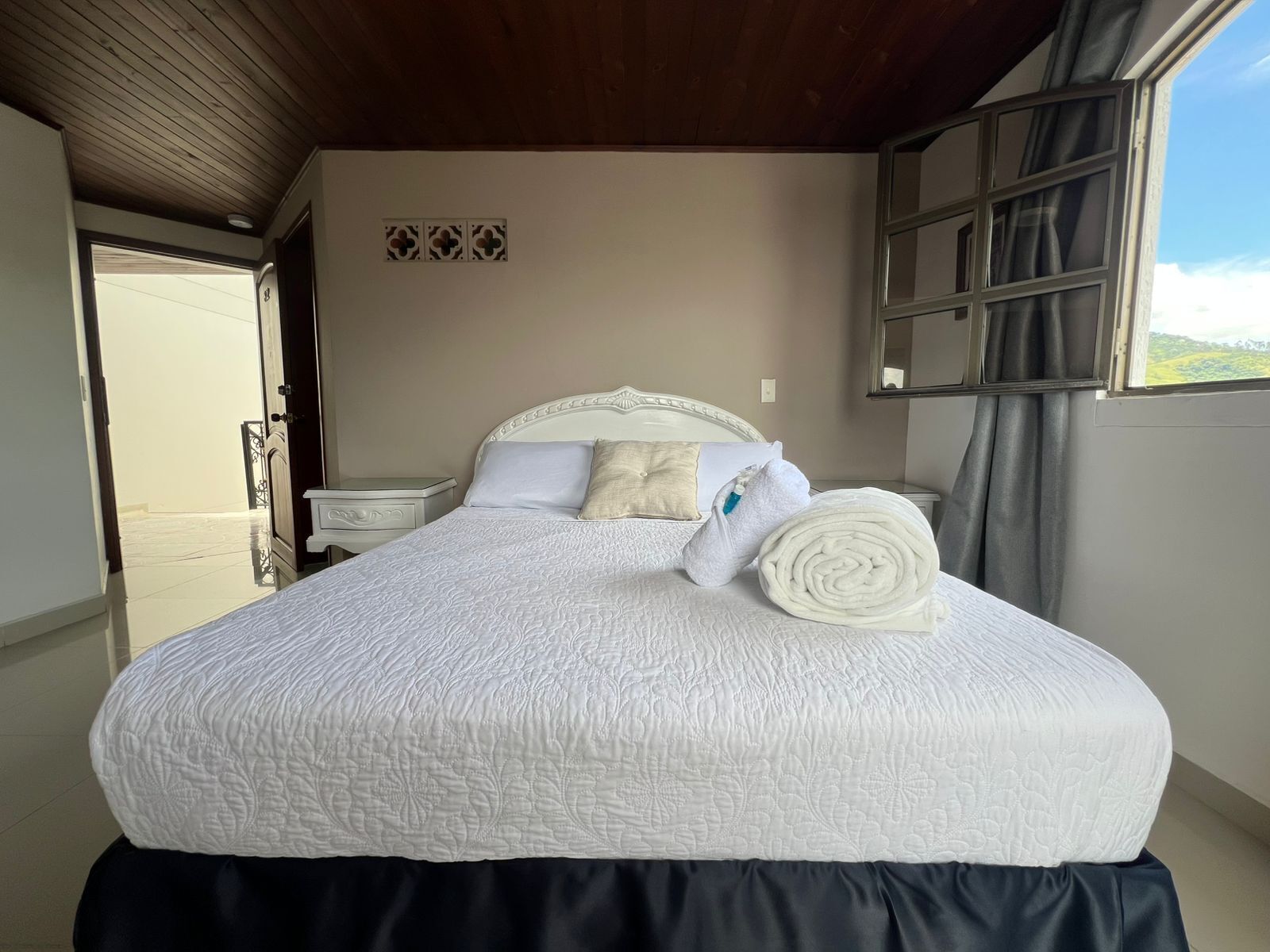 Een bed met een witte sprei, een opgerolde handdoek en een nachtkastje met een lamp.