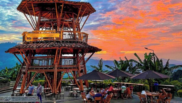 A multi-level wooden structure with outdoor seating and tables, surrounded by lush greenery and a scenic view.