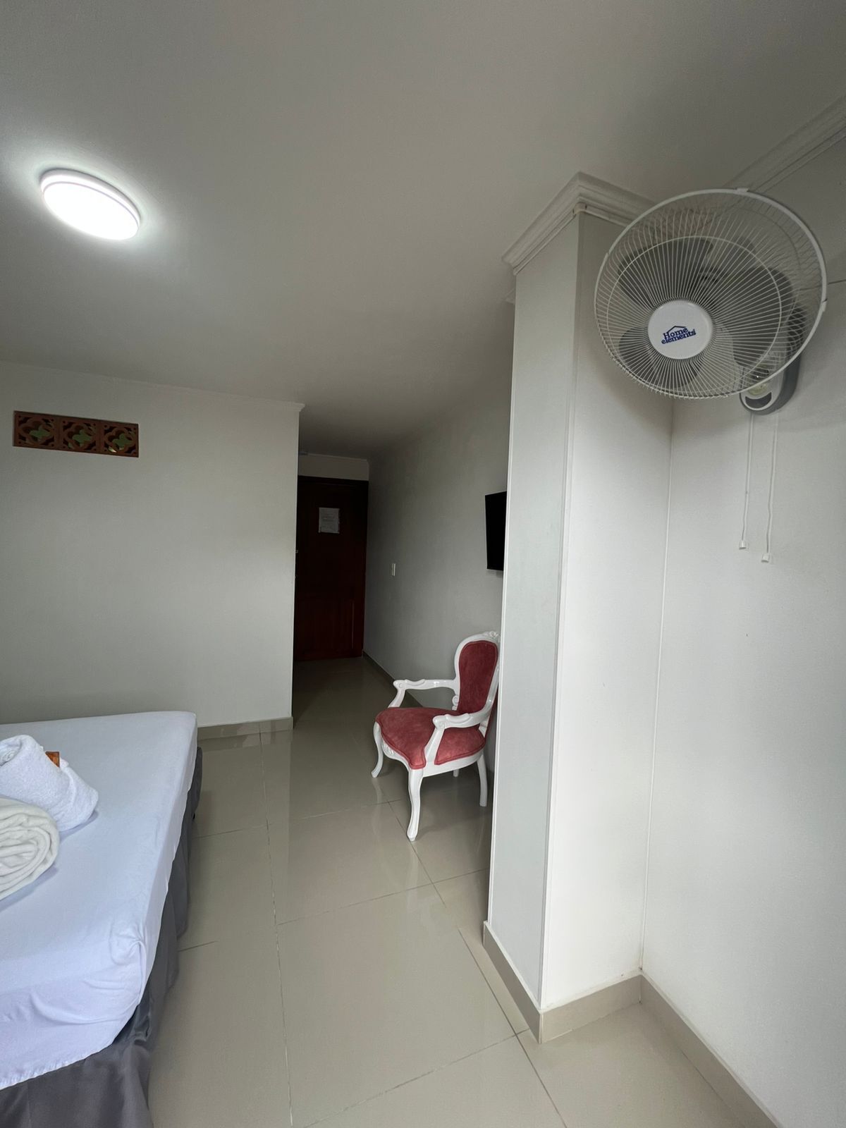 A hotel room with a bed, a chair, a standing fan, and a door leading to another room.