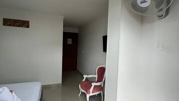 A hotel room with a bed, a chair, a standing fan, and a door leading to another room.