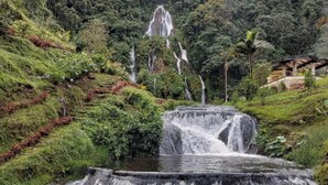 A waterfall cascading over multiple tiers of rock, surrounded by lush greenery and a forested hillside.