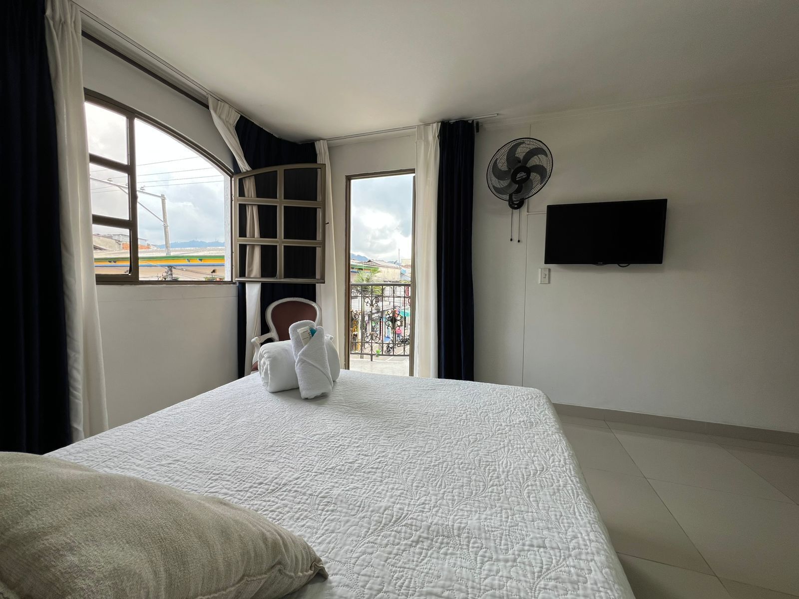 A hotel room with a bed, a chair, a balcony with a view, a fan, and a TV.