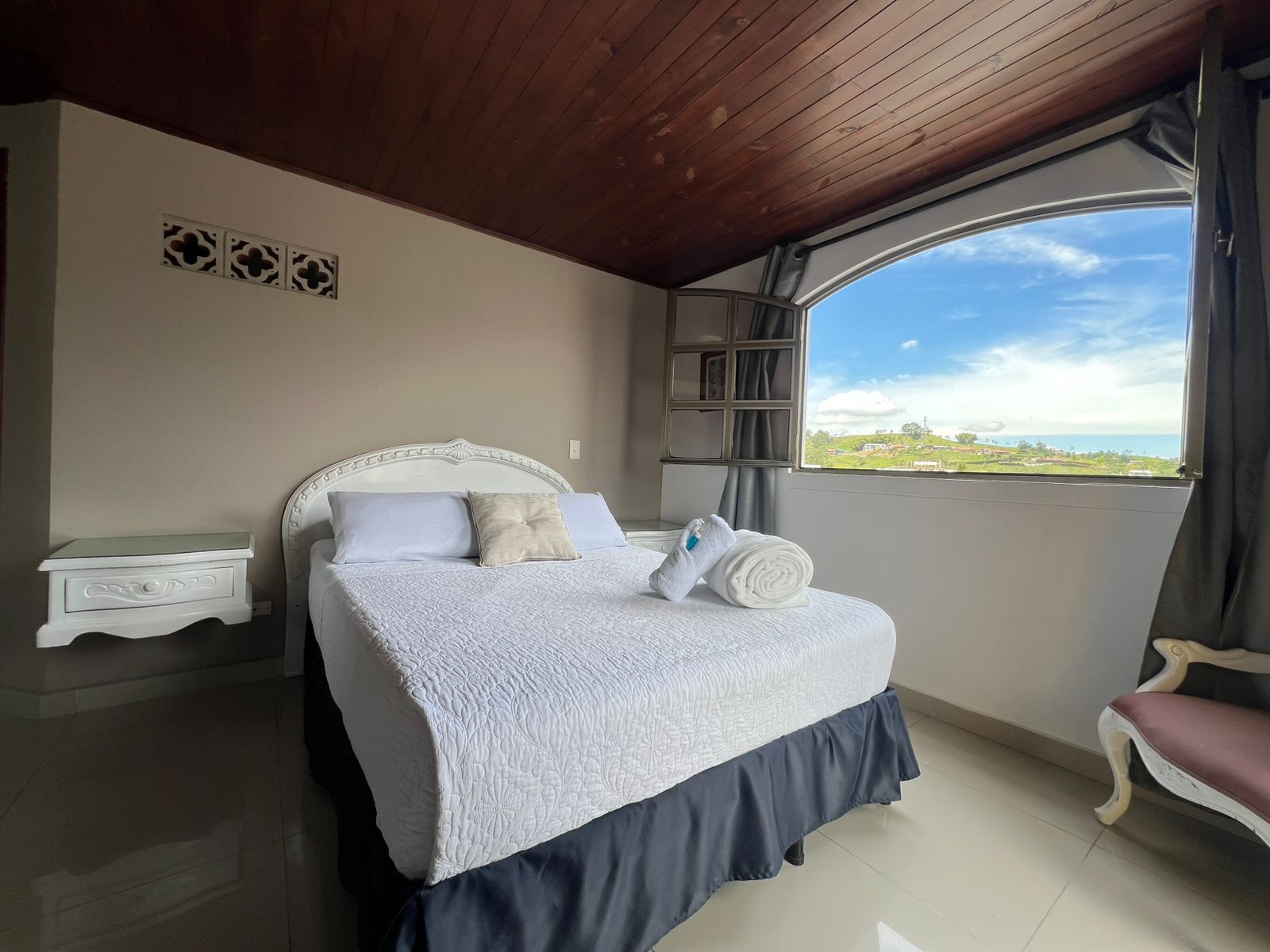A bedroom with a bed, a nightstand, a chair, and a large window with a view.