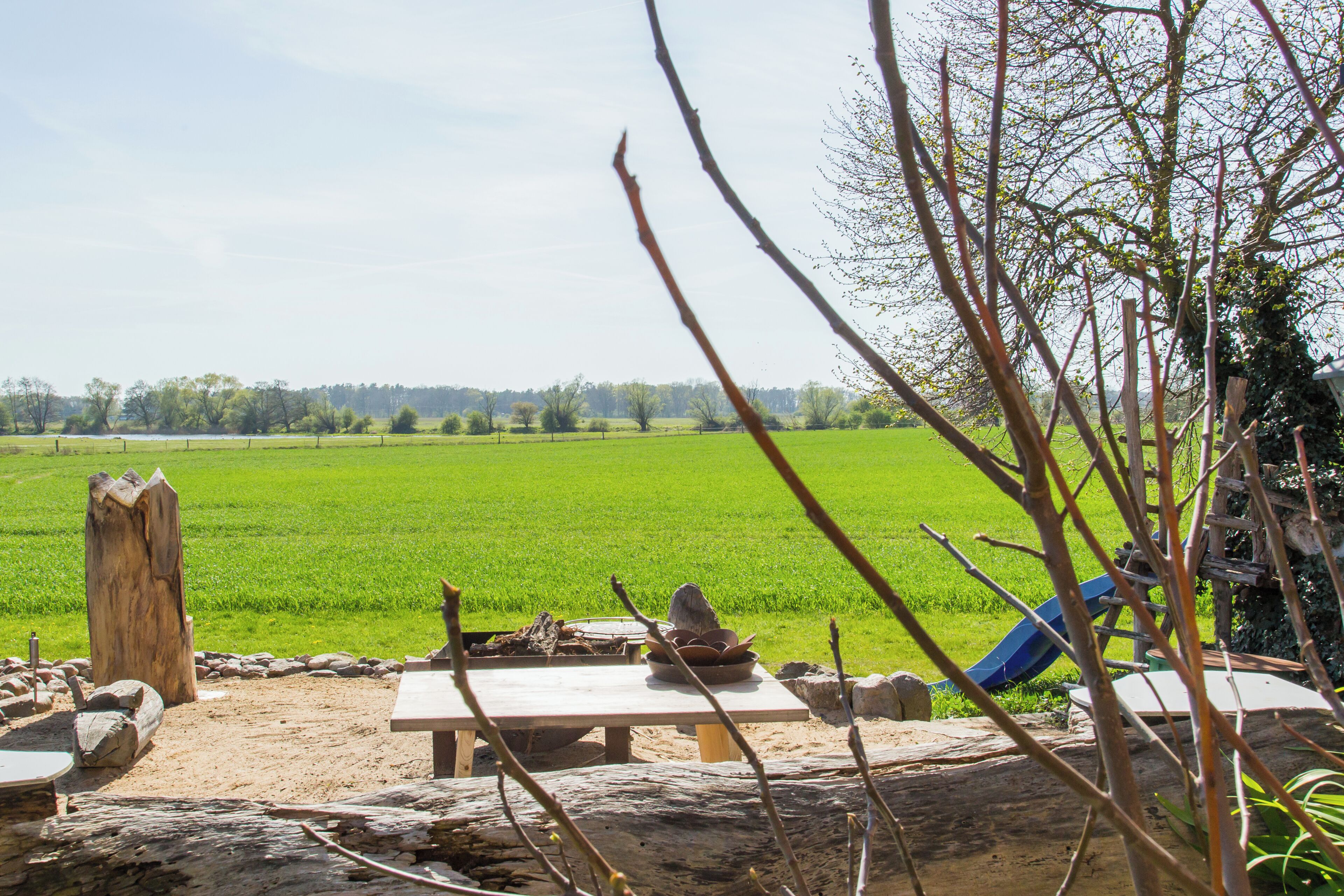 A rustic outdoor dining area with a table and benches, surrounded by a green field and trees.