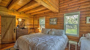 A log cabin bedroom with a large bed, a wooden dresser, a chair, and two windows with blinds. - Log Cabin Home  3 Private Acres (Wimberley)