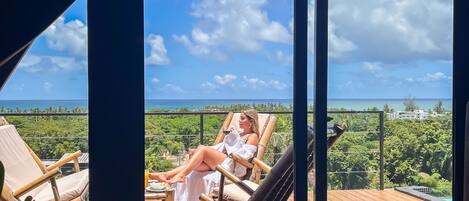 A balcony with a person relaxing on a chair, a table with a drink, and a view of the ocean.