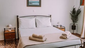 A neatly made bed with white linens, a folded towel, and a framed picture above it. There are two bedside tables with lamps, a potted plant, and a painting on the wall. - 'Stoa Apartment' at Rethymno center (Rethymno)