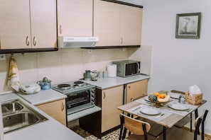A compact kitchen with a stove, microwave, and a sink. There is a dining table set with plates and a basket of fruit. - 'Stoa Apartment' at Rethymno center (Rethymno)