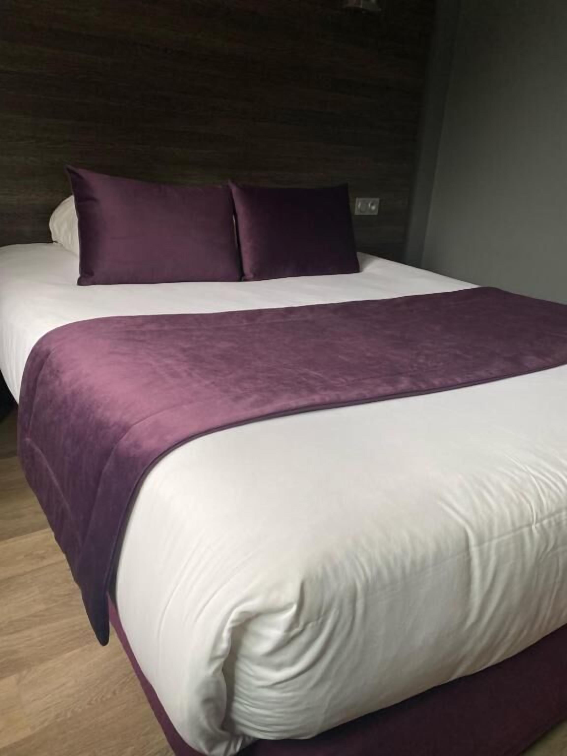 Un lit avec une literie blanche et violette, un plancher en bois et un mur foncé.