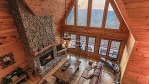 A log cabin interior with a stone fireplace, wooden floors, leather sofas, and large windows offering mountain views. - Blue Ridge Overlook Lodge | Blue Ridge, GA (Cherry Log)