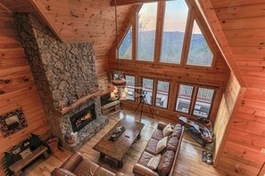 Ein rustikales Blockhaus-Interieur mit Kamin aus Stein, Holzböden, Ledersofas und großen Fenstern mit Blick auf die Berge.