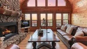 A log cabin interior with a stone fireplace, a wooden table, a leather sofa, and a large window offering a view. - Blue Ridge Overlook Lodge | Blue Ridge, GA (Cherry Log)