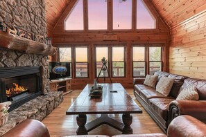 Ein Blockhaushaus-Interieur mit Kamin aus Stein, einem Holztisch, einem Ledersofa und einem großen Fenster mit Blick hinaus.