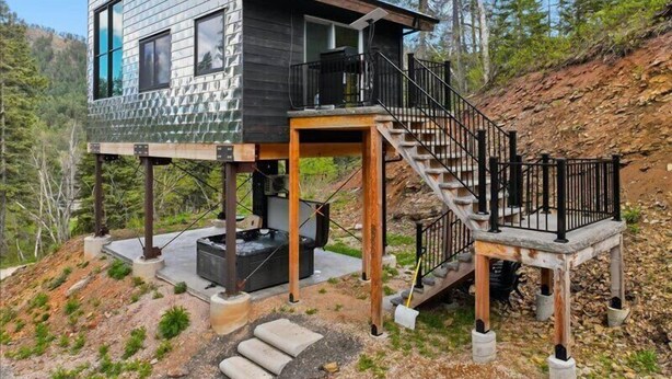 A modern cabin with a wooden deck, a hot tub, and a staircase leading to a balcony.