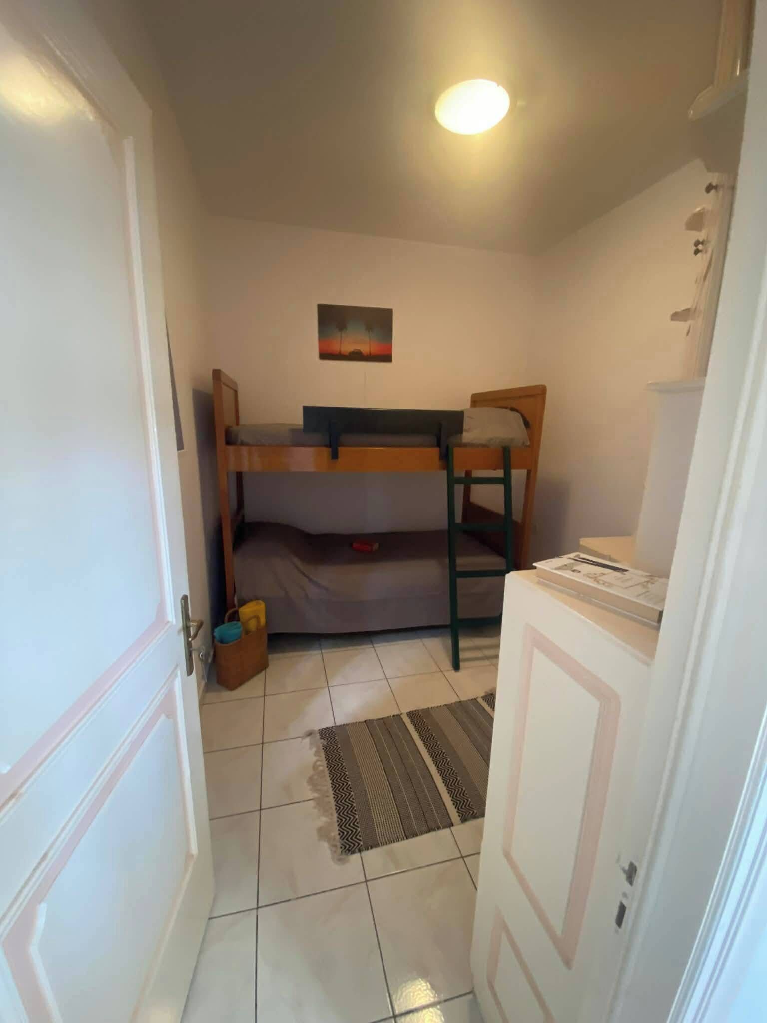 A small, compact room with a bunk bed, a single chair, a small table, and a door.