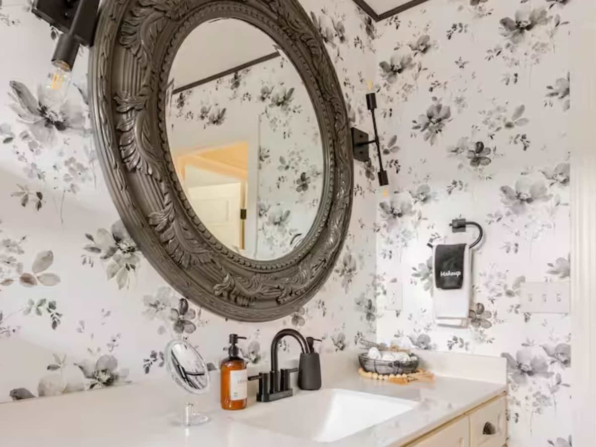 Ein Badezimmer mit rundem Spiegel, floraler Tapete, einem weißen Waschbecken und einem Handtuchhalter.