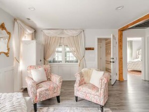 Une pièce avec deux fauteuils à motifs, une grande fenêtre avec des rideaux, un plancher en bois et une porte donnant sur une autre pièce.