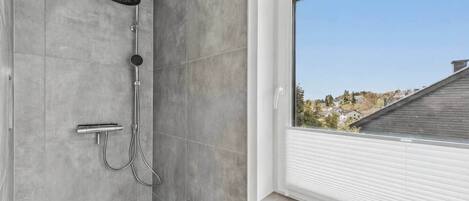 A shower with a handheld showerhead and a wall-mounted faucet, a window with blinds, and a view of a residential area.