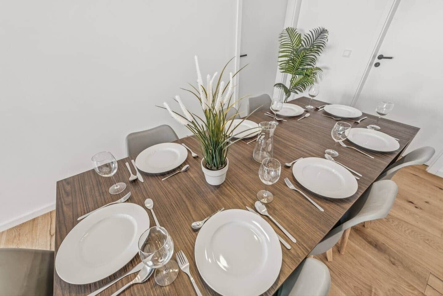 Ein Esstisch für sechs Personen, gedeckt mit weißen Tellern, Besteck und Gläsern, mit einem Blumentopf und einer Karaffe.
