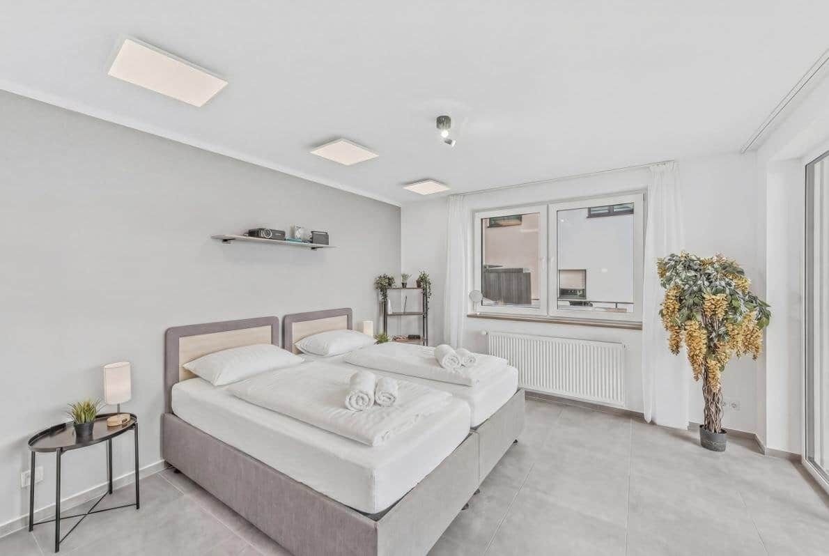 Ein modernes Schlafzimmer mit Doppelbett, Nachttischen, Heizkörper, einem fenster mit Vorhängen und einer Topfpflanze.